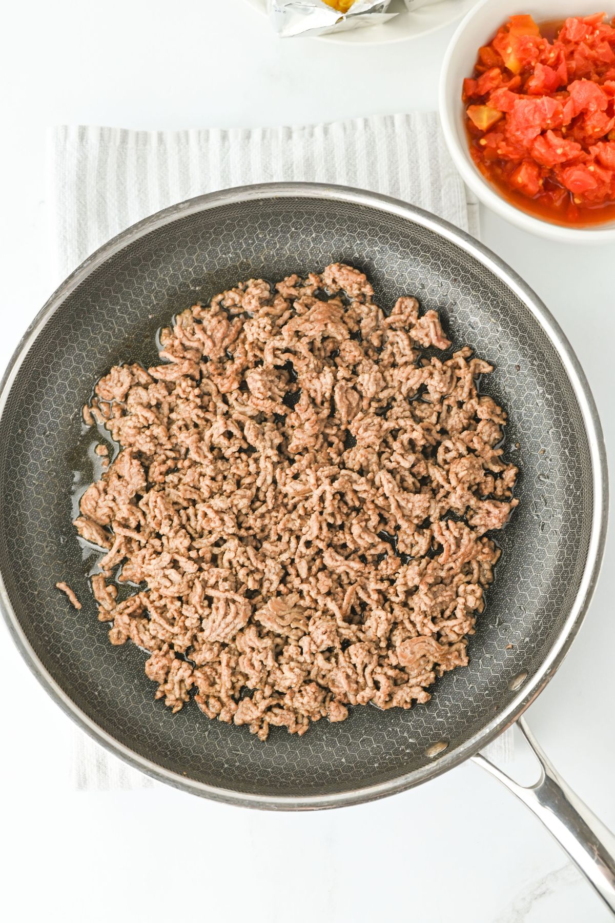 Cooked ground beef in a frying pan on a striped white towel, with a bowl of diced tomatoes visible in the corner of the image.
