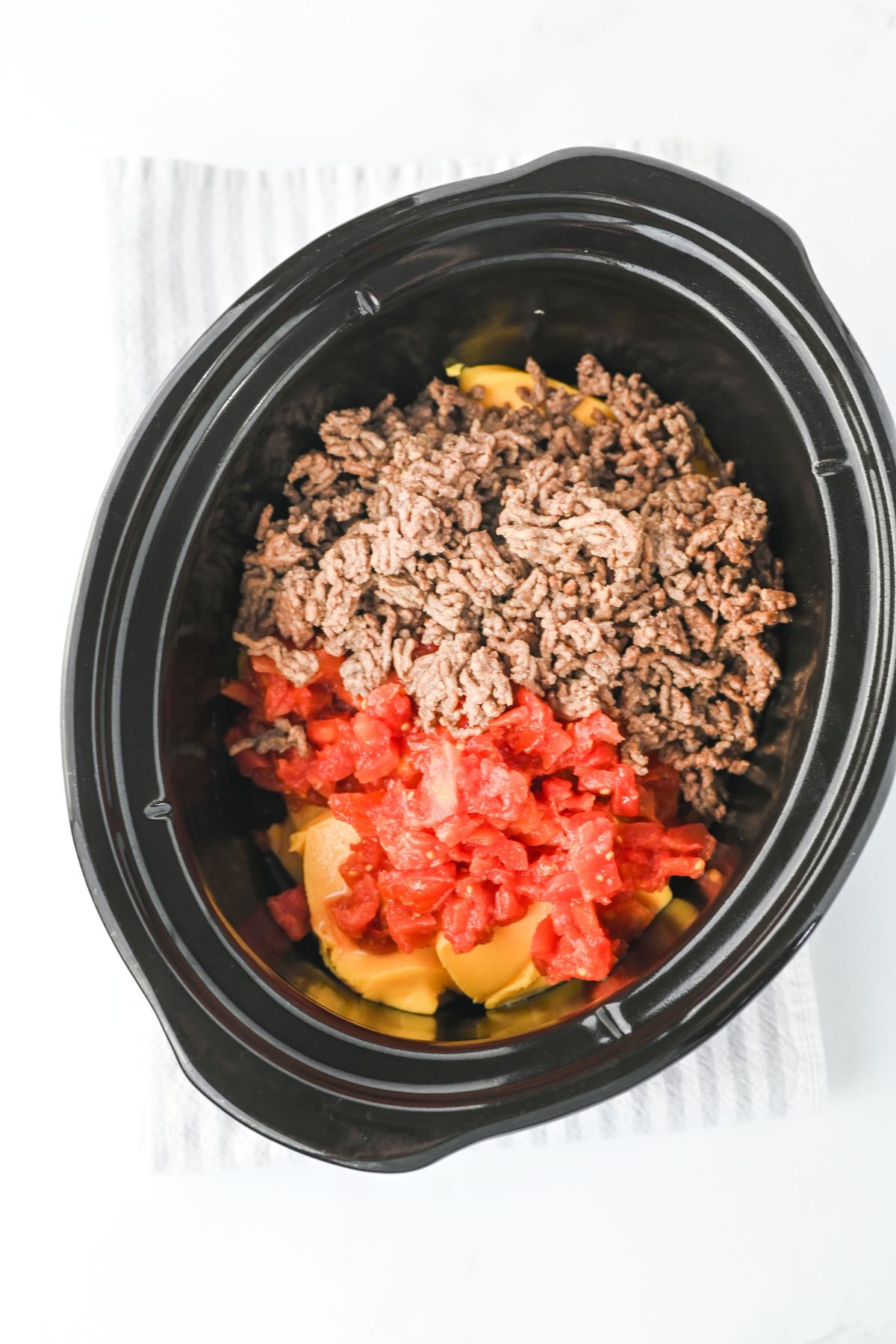 A black slow cooker containing browned ground beef, chopped tomatoes, and sliced bell peppers sits on a white and gray striped cloth. The ingredients are not yet mixed together.
