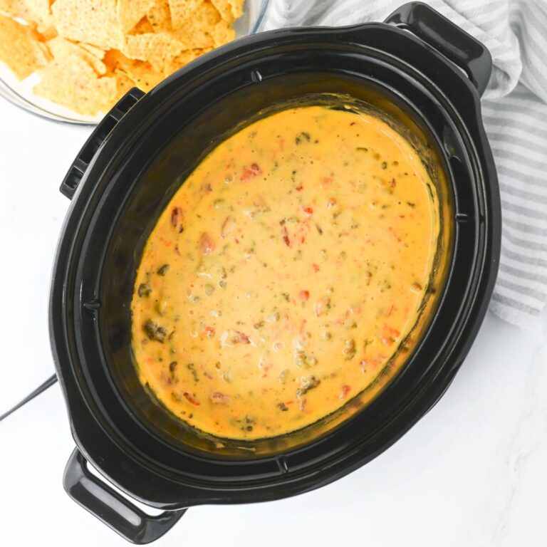 A black slow cooker filled with melted queso dip containing visible chunks of tomato and green herbs. A bowl of tortilla chips is partially visible in the background.