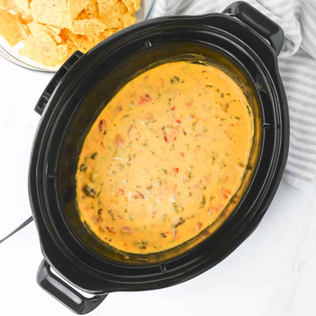 A black slow cooker filled with melted queso dip containing visible chunks of tomato and green herbs. A bowl of tortilla chips is partially visible in the background.