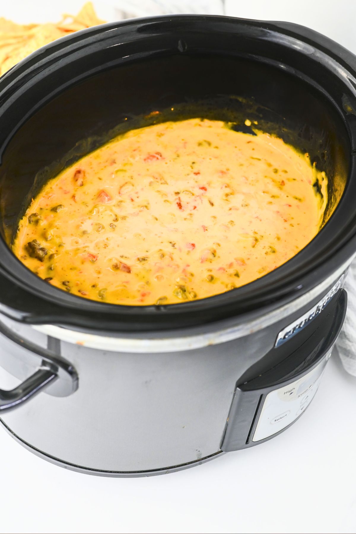 A slow cooker filled with a creamy, orange-yellow cheese dip mixed with visible chunks of tomatoes and green peppers. Some tortilla chips are partially visible in the background.