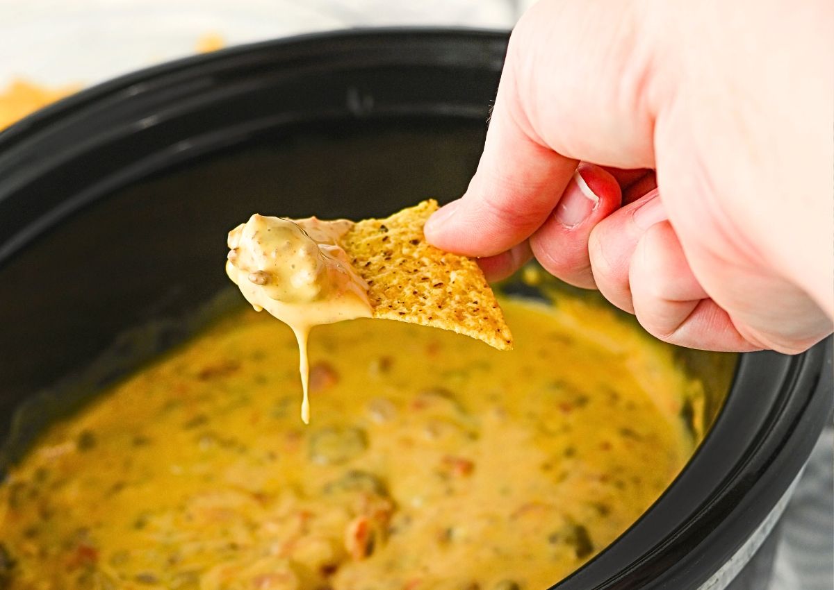 A hand holds a tortilla chip dipped in a thick, cheesy dip with visible chunks, above a slow cooker filled with the same dip.