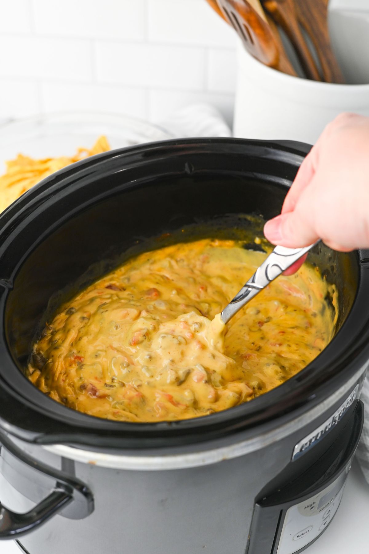 A hand stirs a thick, yellow cheese dip with visible chunks of tomatoes and beans inside a black slow cooker. Tortilla chips and kitchen utensils are visible in the background.