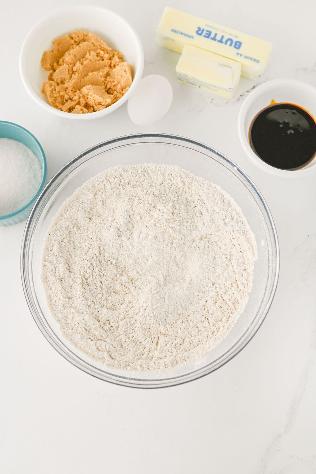 A glass bowl filled with flour mixture sits beside small bowls of brown sugar, white sugar, a stick of butter, an egg, and a dish of dark liquid, all arranged on a white surface.