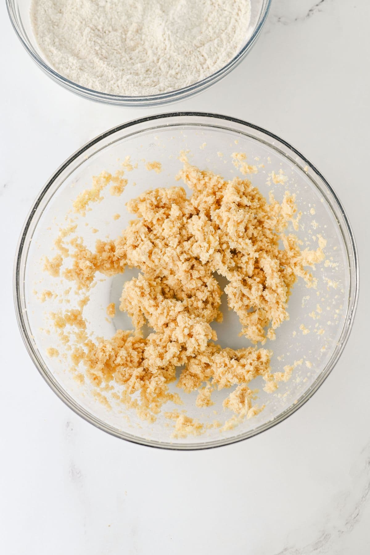 A glass bowl containing a creamed mixture of butter and sugar, with a second bowl of flour visible in the background on a white surface.