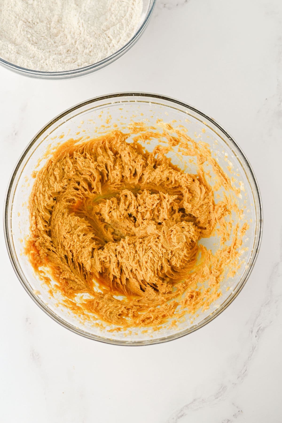 A glass bowl containing creamed butter and sugar sits on a white surface. Another glass bowl with flour is partially visible at the top of the image.