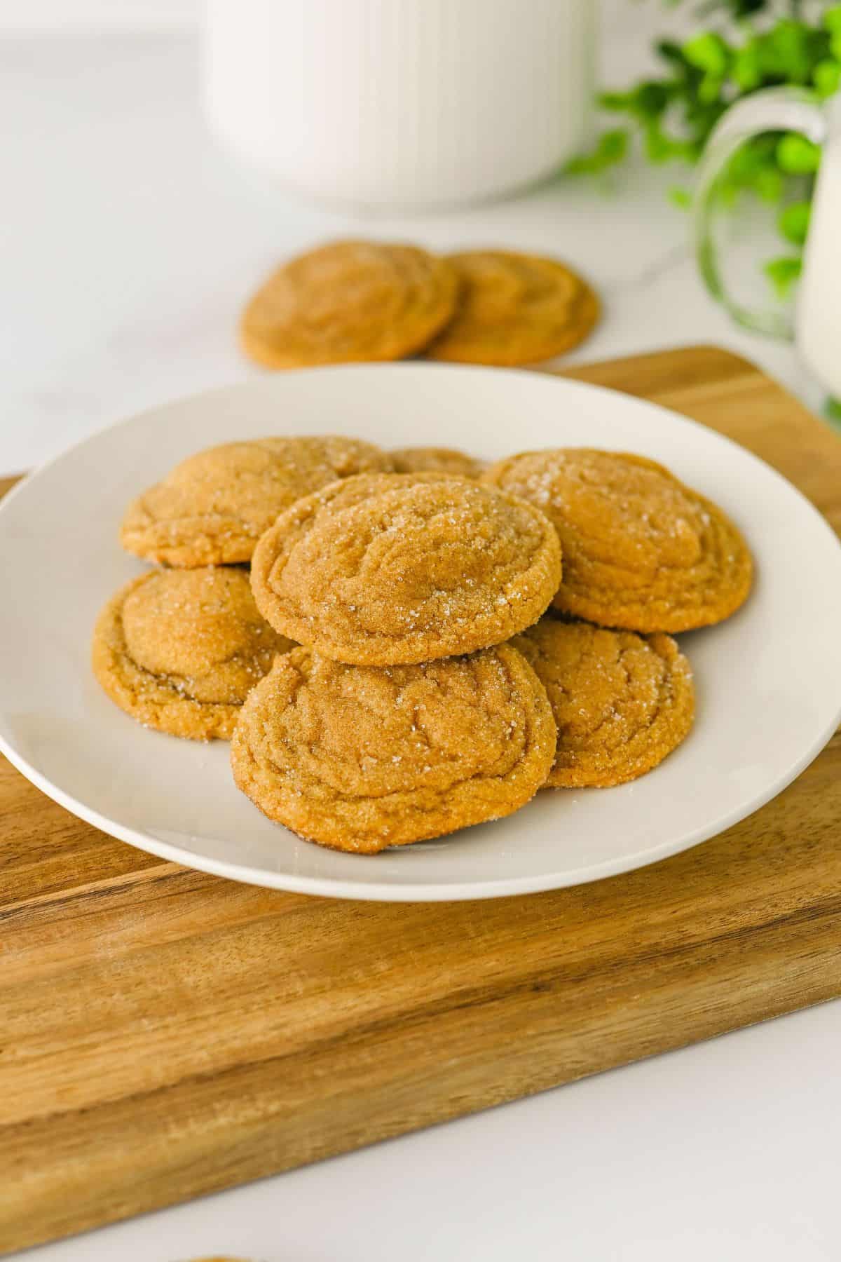 A white plate with a stack of sugar-coated cookies sits on a wooden surface. More cookies, a jar, a green plant, and a glass of milk are in the background.