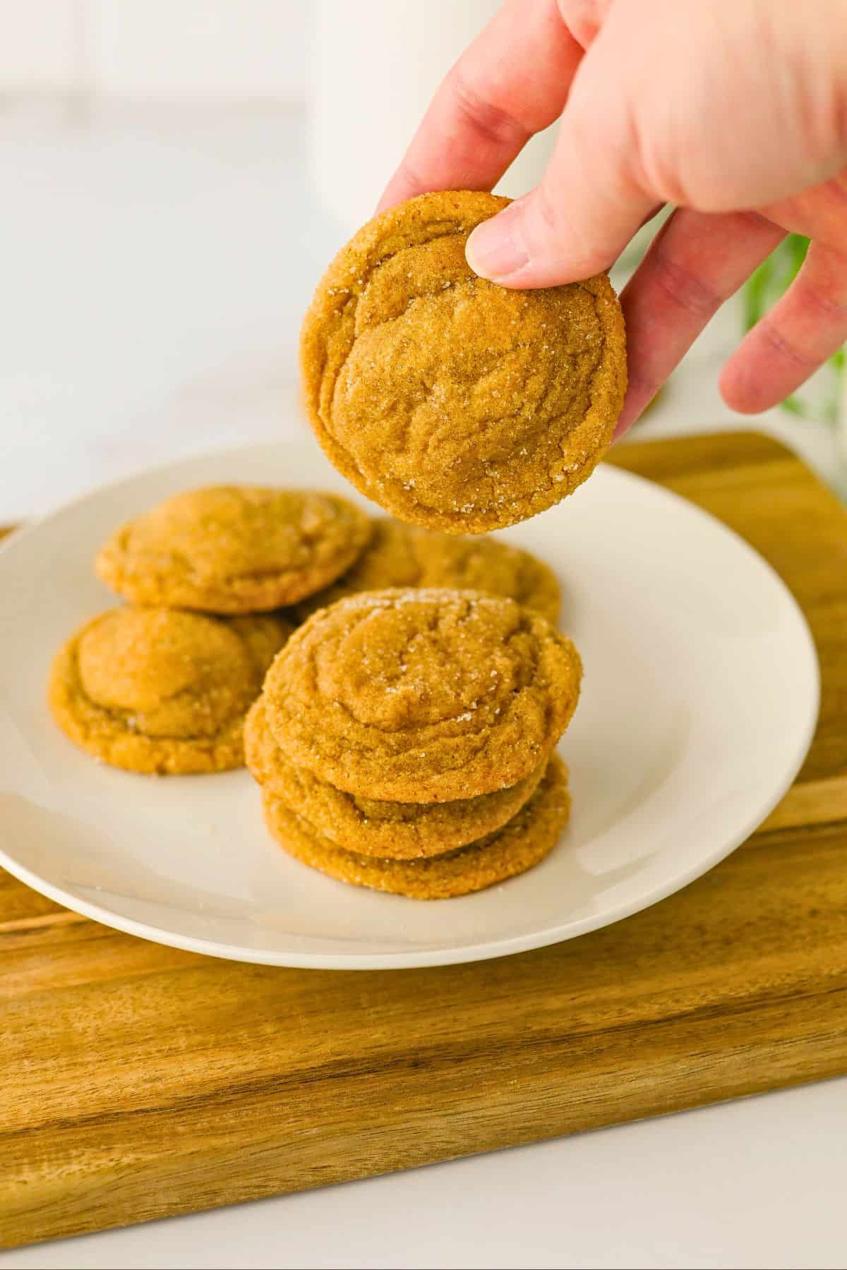 A hand holds a round brown cookie above a white plate with a stack of similar cookies, all on a wooden board.
