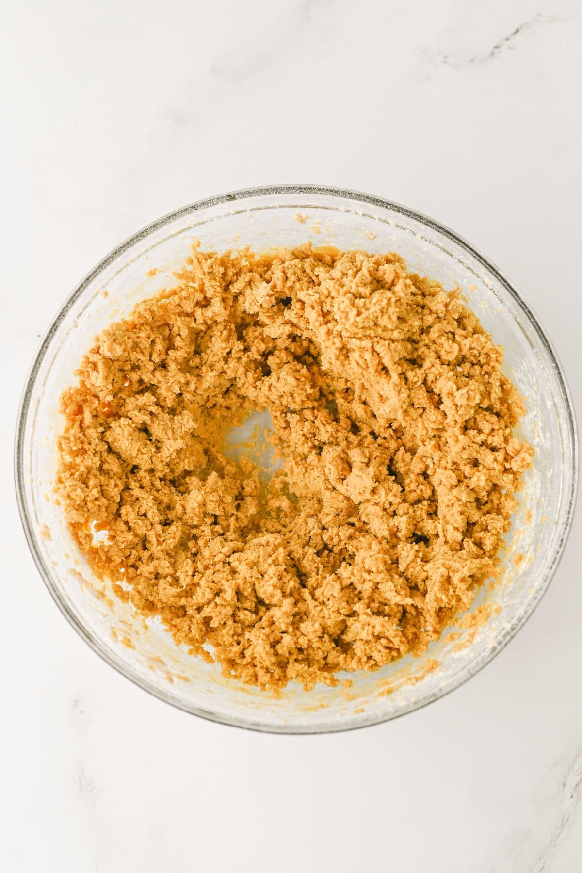A glass mixing bowl on a white surface filled with a thick, light brown cookie dough mixture, with a small well or indentation in the center.