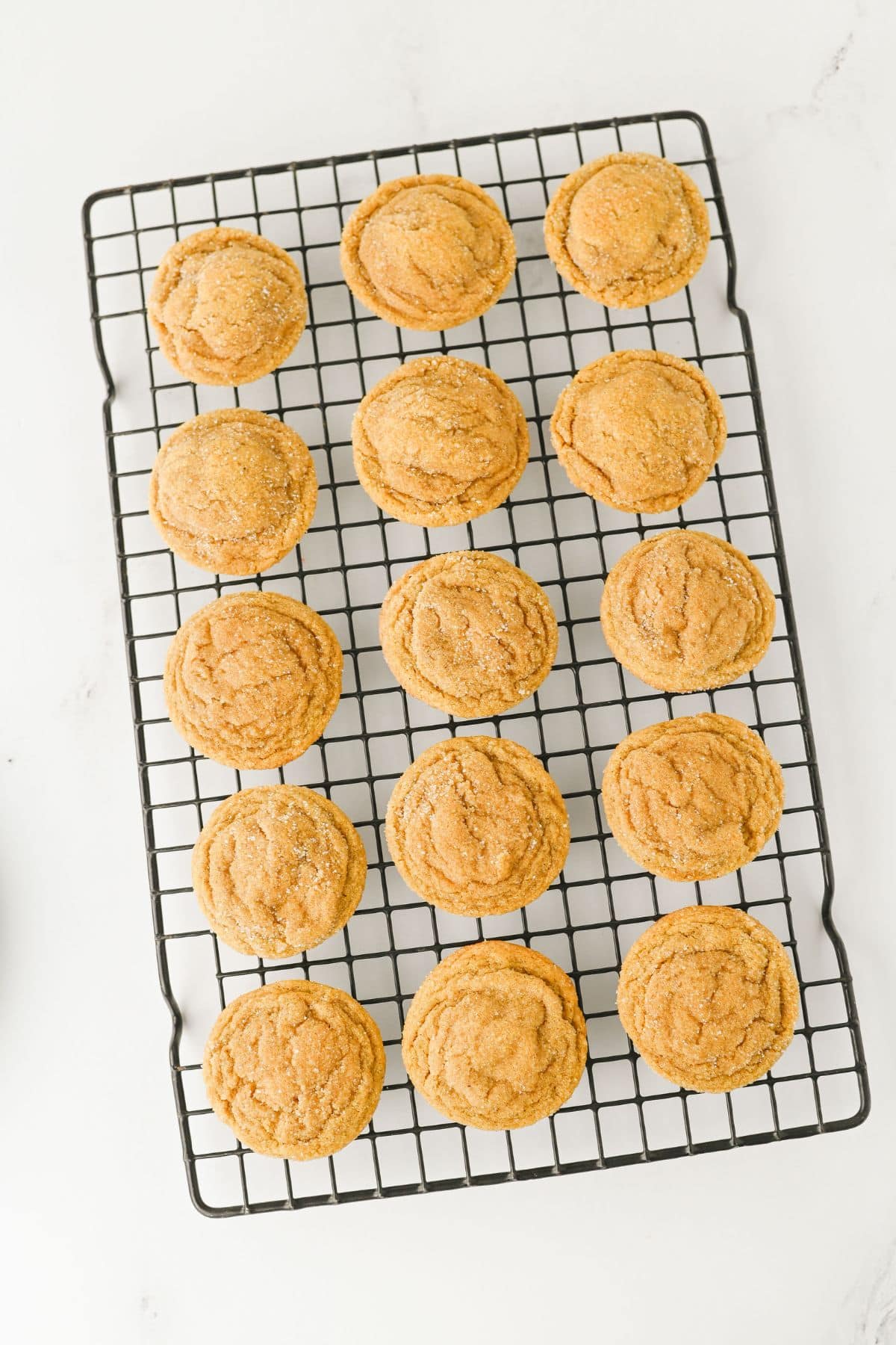 A cooling rack with fifteen evenly spaced, golden-brown cookies arranged in three rows on a white surface.