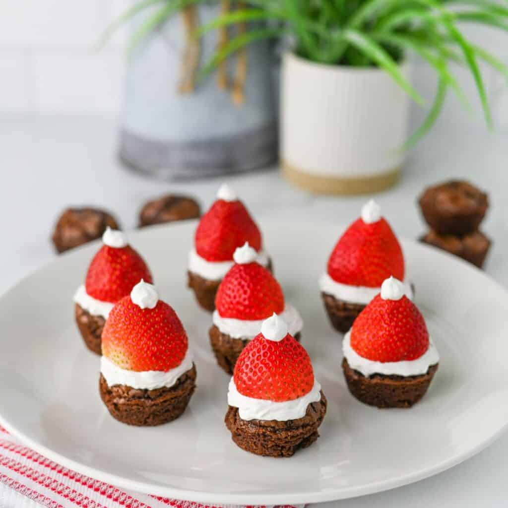 A white plate holds several Santa Hat Brownie Bites, each topped with a layer of white frosting, a whole strawberry, and a small dollop of frosting to resemble Santa hats. A plant and blurred objects are in the background.