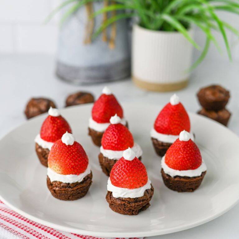 A white plate holds several Santa Hat Brownie Bites, each topped with a layer of white frosting, a whole strawberry, and a small dollop of frosting to resemble Santa hats. A plant and blurred objects are in the background.