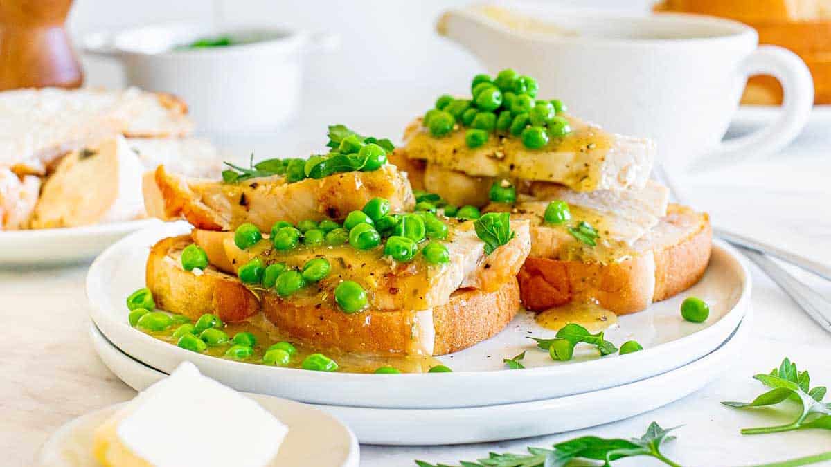 Slices of roasted turkey on toasted bread are topped with green peas, gravy, and parsley, creating one of the 18 Amazing Sandwiches, served on a white plate with butter and additional food items in the background.