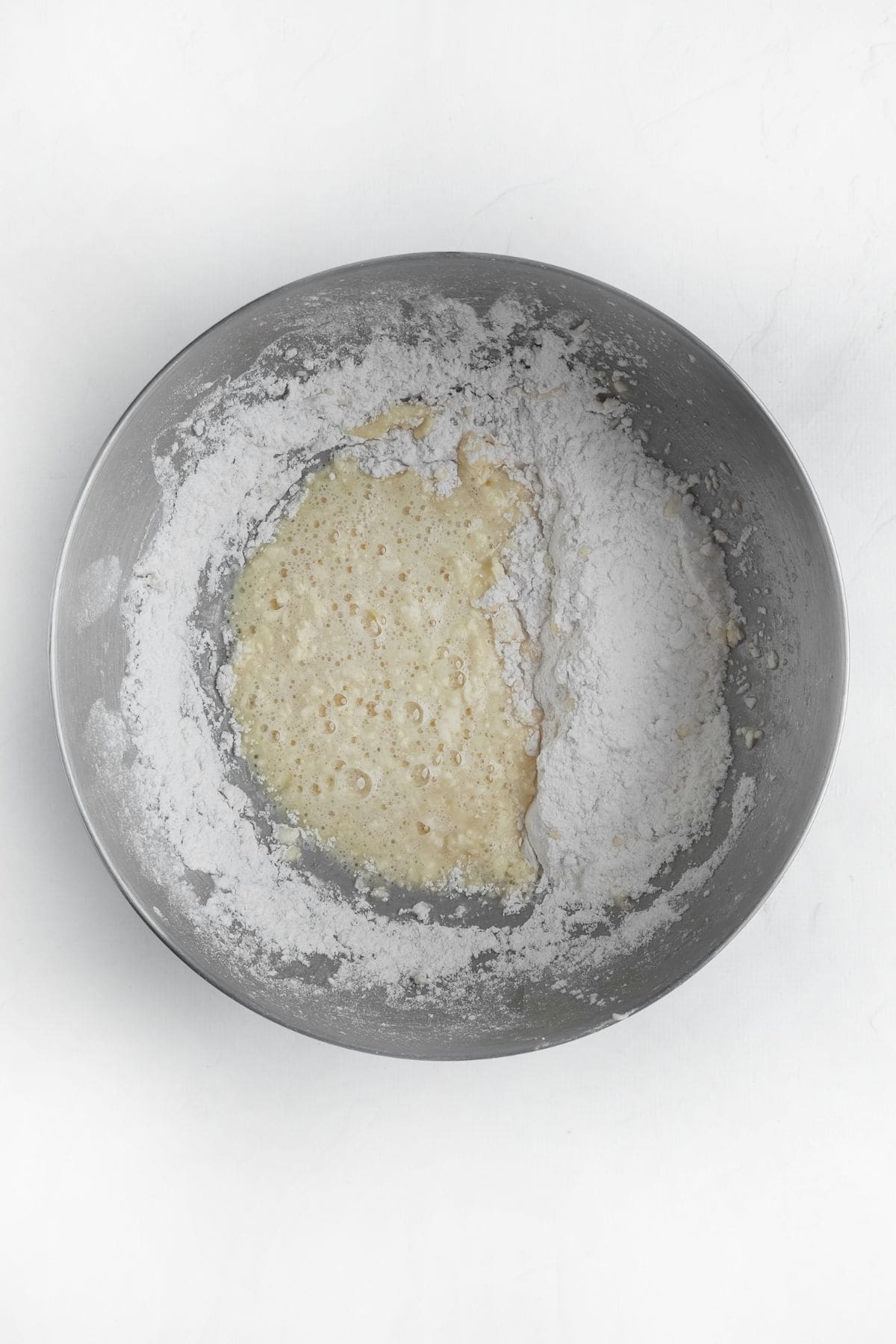 A metal mixing bowl containing a mixture of flour and a bubbly liquid batter on a white surface. The ingredients are partially combined, showing distinct wet and dry areas.