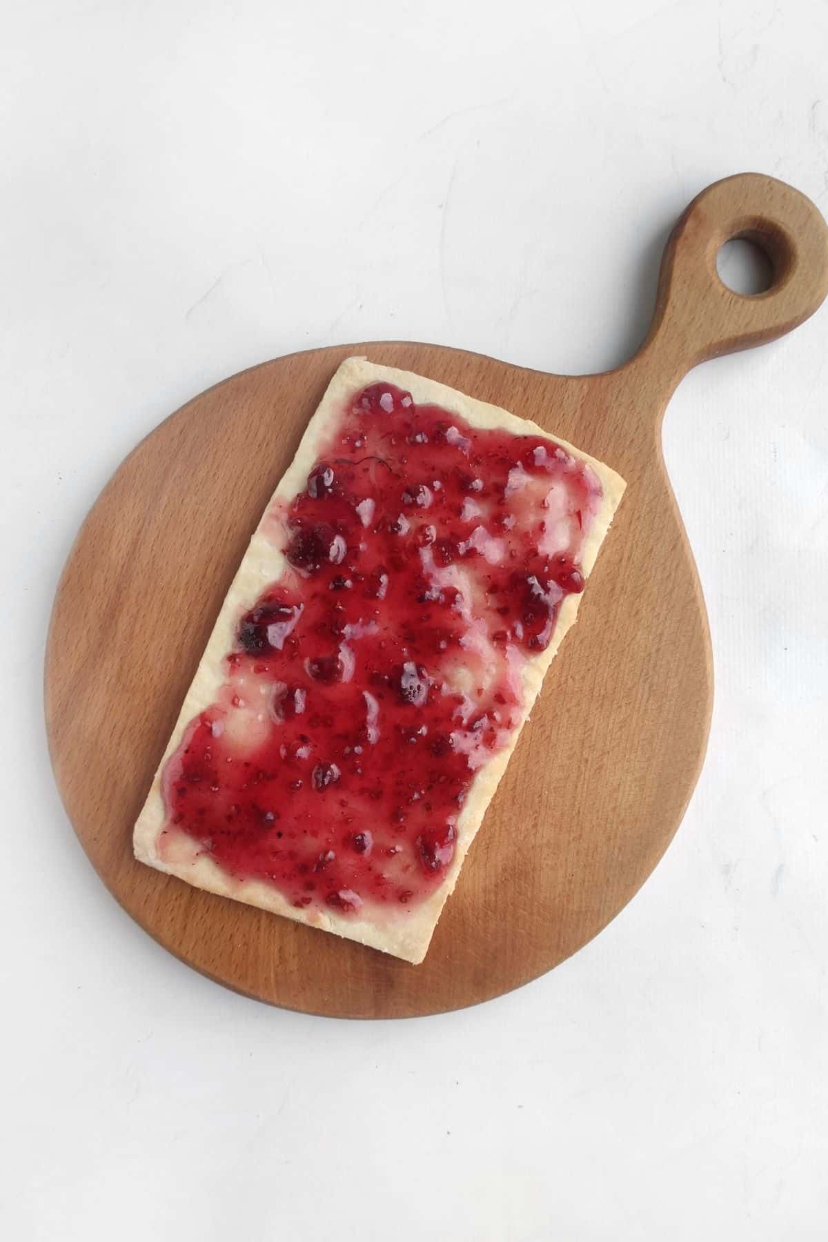 A rectangular piece of bread with red jam spread on top, placed on a round wooden cutting board with a handle, set against a plain white background.
