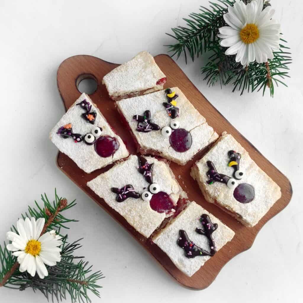 A wooden board holds five rectangular pieces of powdered sugar-topped pastry decorated with reindeer faces. The board is surrounded by pine sprigs and white daisy flowers on a white surface.