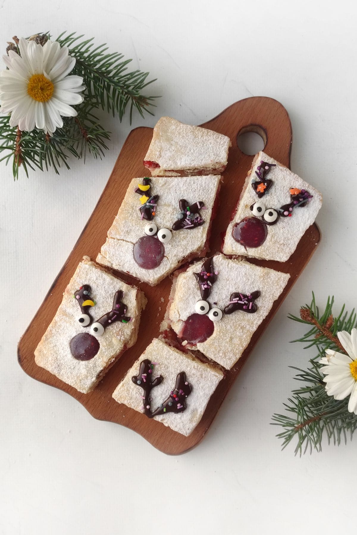 Rectangular pieces of cake with powdered sugar are decorated with icing and candies to look like reindeer faces. The cakes are on a wooden board with pine sprigs and white flowers beside them.