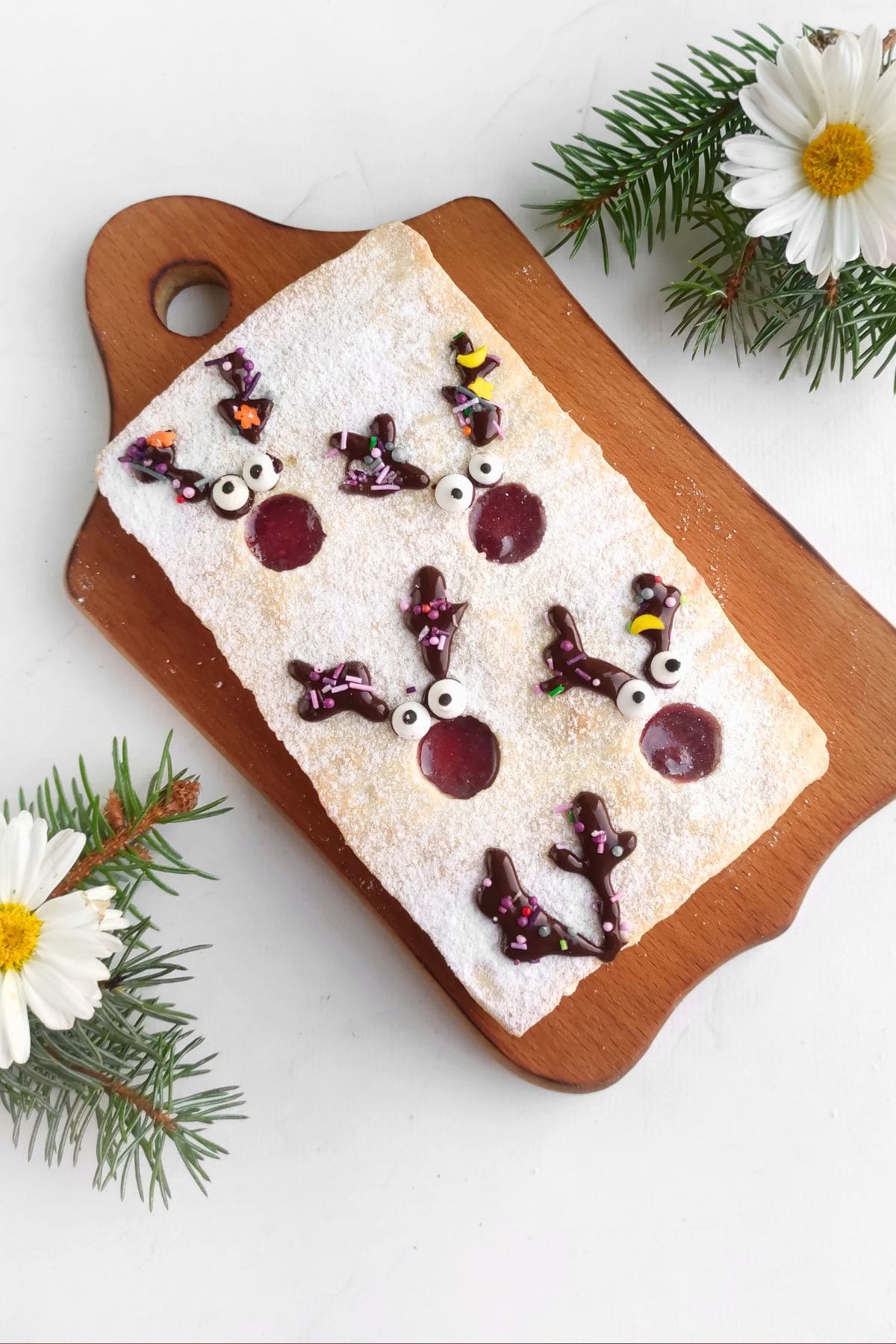 A rectangular pastry dusted with powdered sugar sits on a wooden board. It is decorated with chocolate reindeer faces and red jelly circles. Pine branches and white daisies are placed nearby on the white surface.