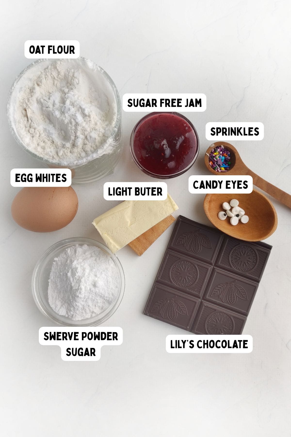 A top-down view of baking ingredients, each labeled: oat flour in a bowl, an egg, powdered sweetener, a stick of light butter, sugar-free jam in a dish, chocolate squares, candy eyes in a small bowl, and colorful sprinkles on a spoon.