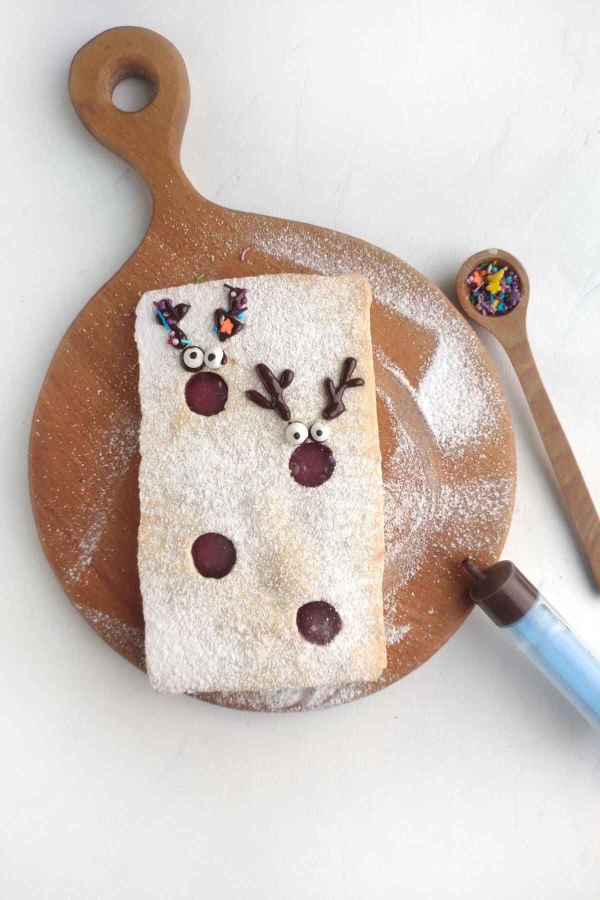 A rectangular pastry dusted with powdered sugar on a wooden board, decorated with two reindeer faces made from chocolate, candy eyes, and sprinkles; a small wooden spoon holds colorful sprinkles beside a blue piping tool.