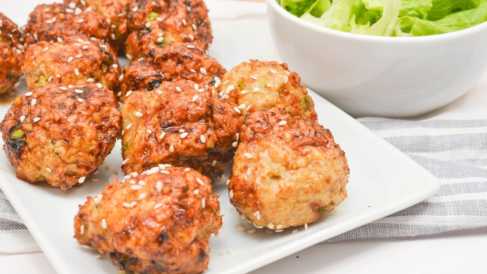 A white rectangular plate with several browned, round falafel balls topped with sesame seeds, next to a white bowl filled with green lettuce leaves. The plate rests on a striped cloth.