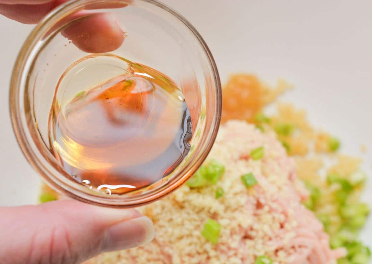 A hand holds a small glass bowl containing a brown liquid above a mixture of breadcrumbs, chopped green onions, and minced ingredients in a white bowl, preparing to pour the liquid in.