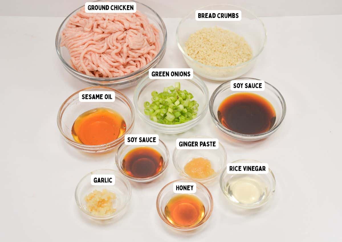 A variety of labeled ingredients in glass bowls on a white surface, including ground chicken, bread crumbs, green onions, soy sauce, sesame oil, ginger paste, rice vinegar, honey, and minced garlic.