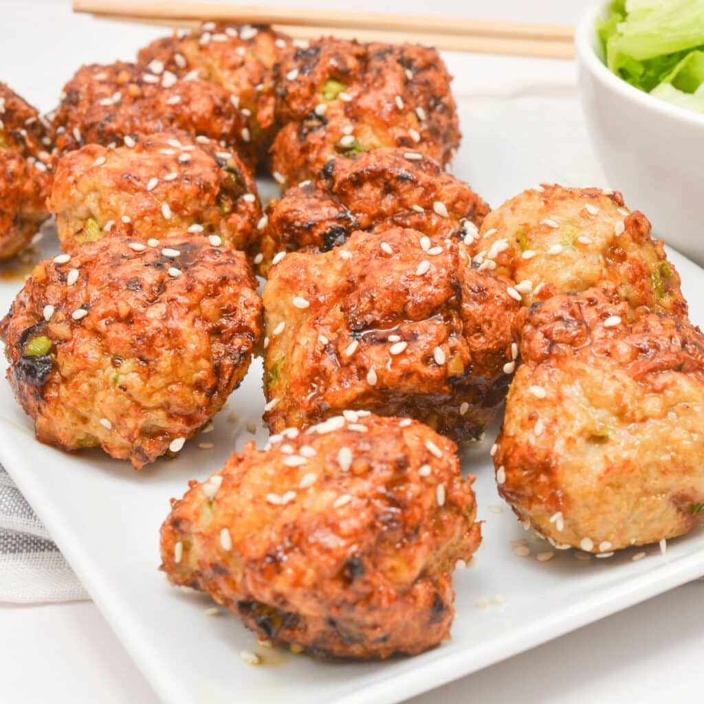A white plate with several golden-brown, round fried meatballs topped with white sesame seeds. A bowl of green salad and a pair of chopsticks are partially visible in the background.