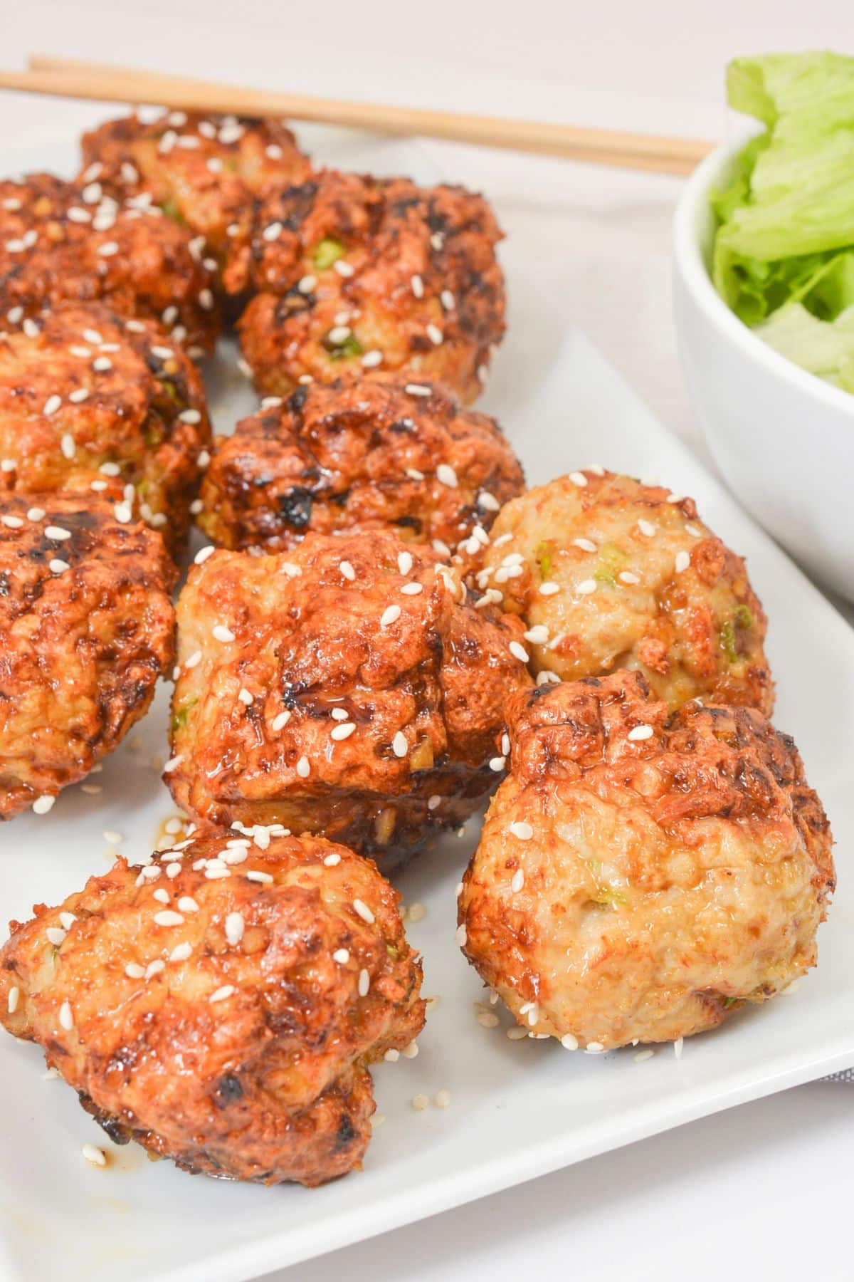 A white rectangular plate with several browned meatballs topped with white sesame seeds. A bowl of leafy green lettuce and a pair of wooden chopsticks are in the background.