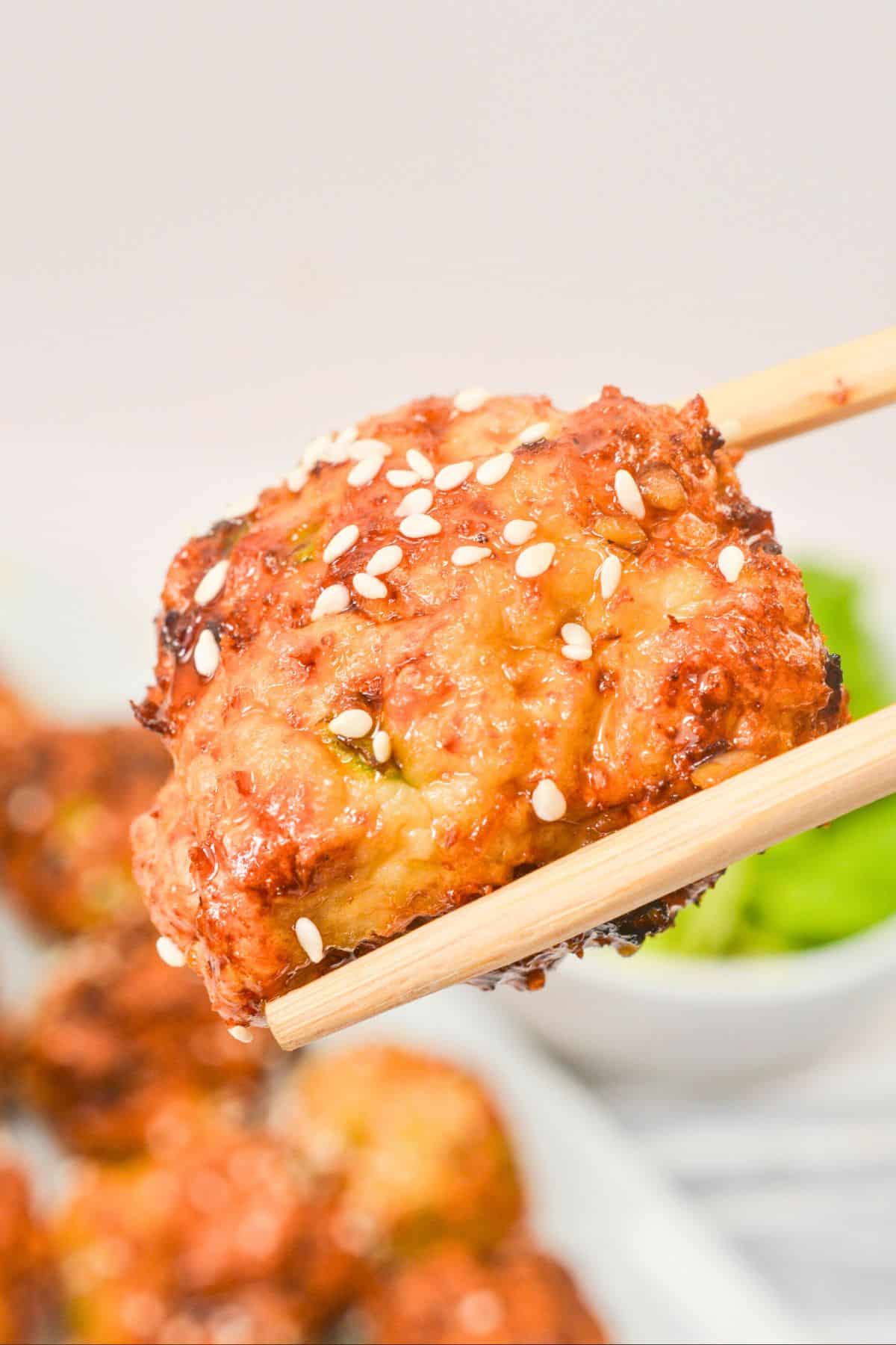 A pair of chopsticks holds a piece of glazed, cooked chicken topped with white sesame seeds. The background is blurred, showing more pieces of similar chicken and a dish with green contents.