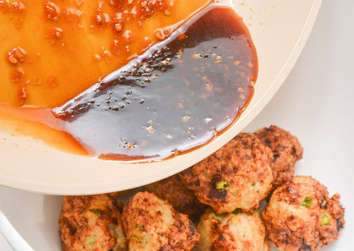 A pan with dark and orange sauce being poured over a bowl of browned meatballs with visible green herbs. The sauce looks glossy and thick, partially covering the meatballs.