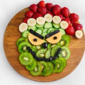 A round wooden board with fruit and vegetable slices arranged to resemble a face, made of strawberries, banana, cucumber, kiwi, and cheese. Black olives and possibly licorice create angry eyebrows and a frown.