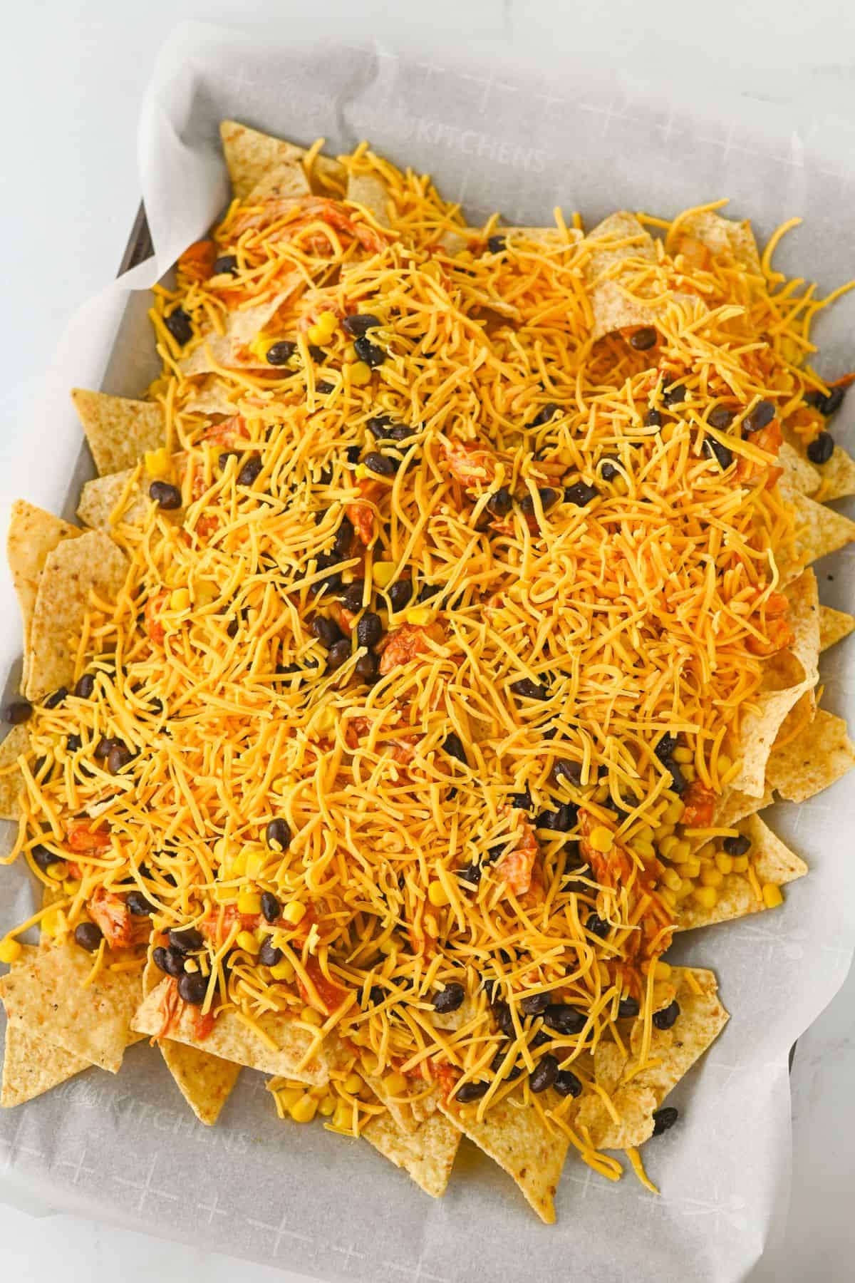 A baking tray lined with parchment paper holding tortilla chips topped with shredded cheese, black beans, corn, and some scattered bits of tomato.