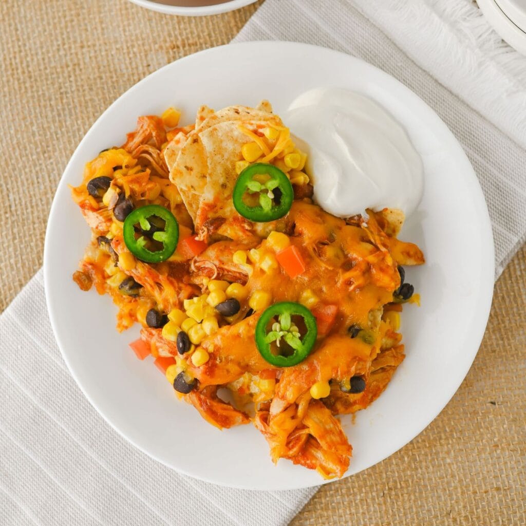 A white plate holds a serving of chicken nachos topped with cheese, black beans, corn, sliced jalapeños, and a side of sour cream. The plate rests on a white napkin over a burlap surface.