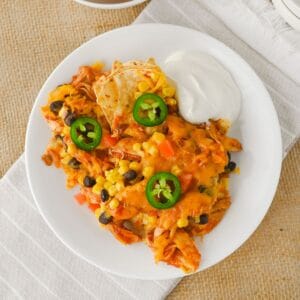 A white plate holds a serving of chicken nachos topped with cheese, black beans, corn, sliced jalapeños, and a side of sour cream. The plate rests on a white napkin over a burlap surface.