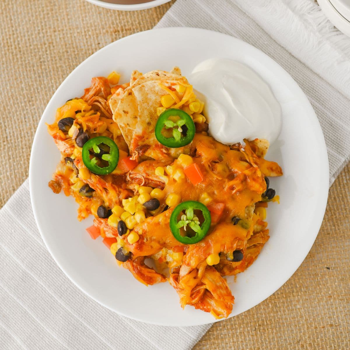 A white plate holds a serving of chicken nachos topped with cheese, black beans, corn, sliced jalapeños, and a side of sour cream. The plate rests on a white napkin over a burlap surface.