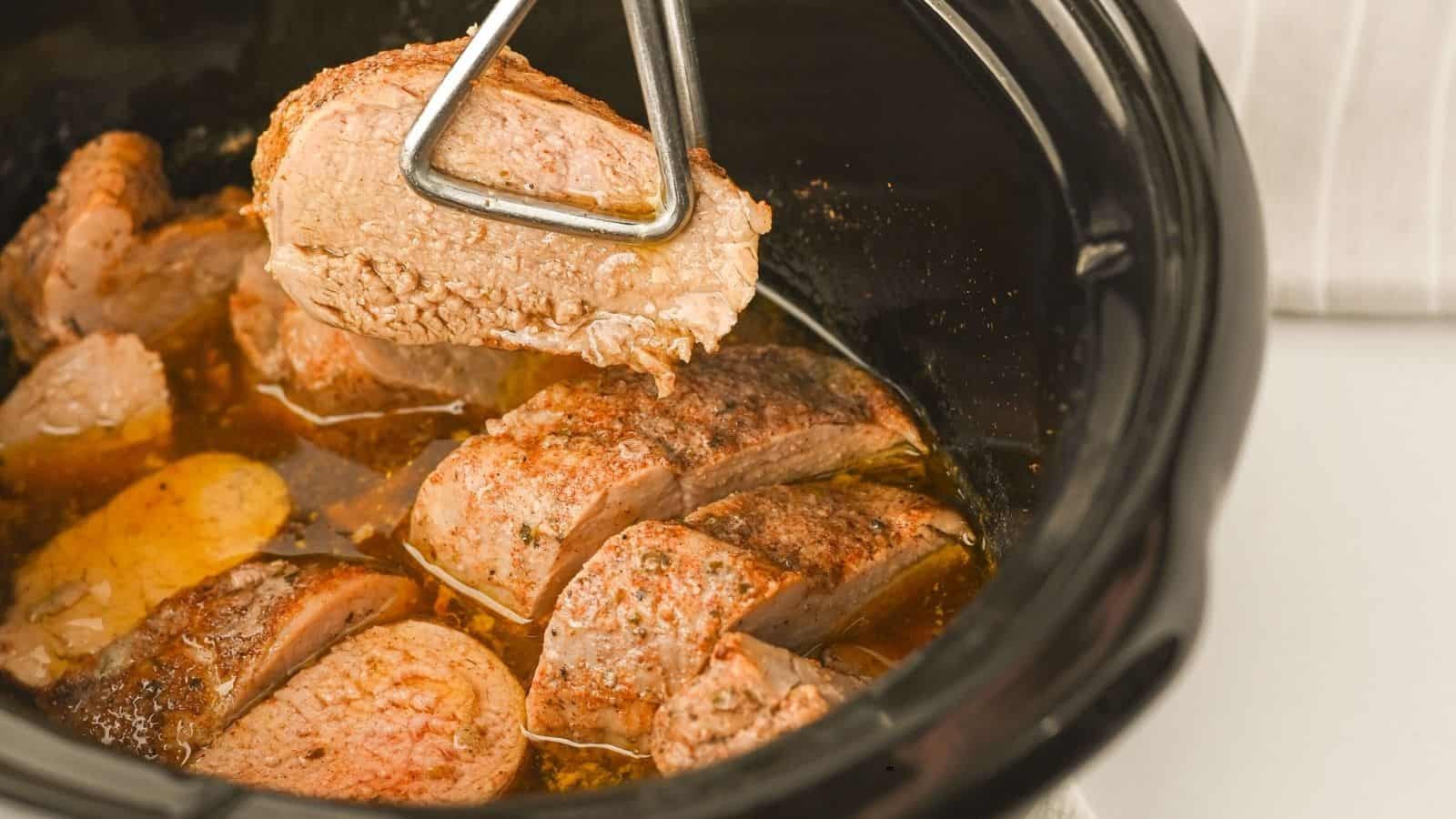 A close-up of sliced, cooked beef in a slow cooker. One piece is being lifted with metal tongs, revealing a browned exterior and moist texture—much like a Crock Pot Pork Tenderloin. Liquid and seasoning are visible at the bottom of the cooker.