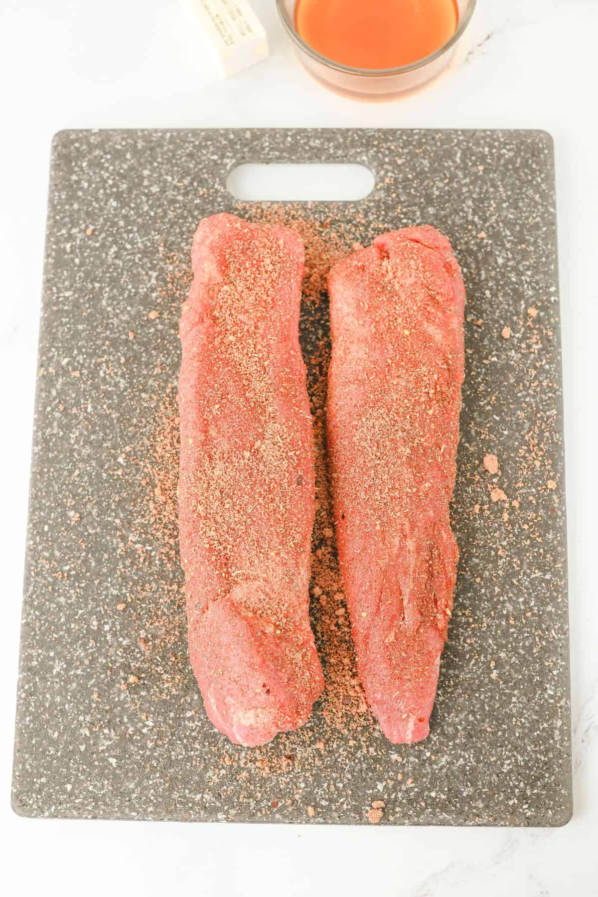 Two raw pork tenderloins coated with dry seasoning rest on a dark gray cutting board, ready for the Crock Pot Pork Tenderloin recipe. A stick of butter and a bowl of orange-brown liquid are partially visible in the top corners.