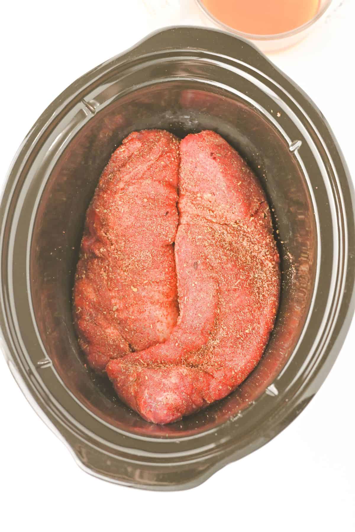Raw, seasoned beef roast sits in a black slow cooker, viewed from above—much like preparing Crock Pot Pork Tenderloin. A container with brown liquid is partially visible in the upper right corner of the image.