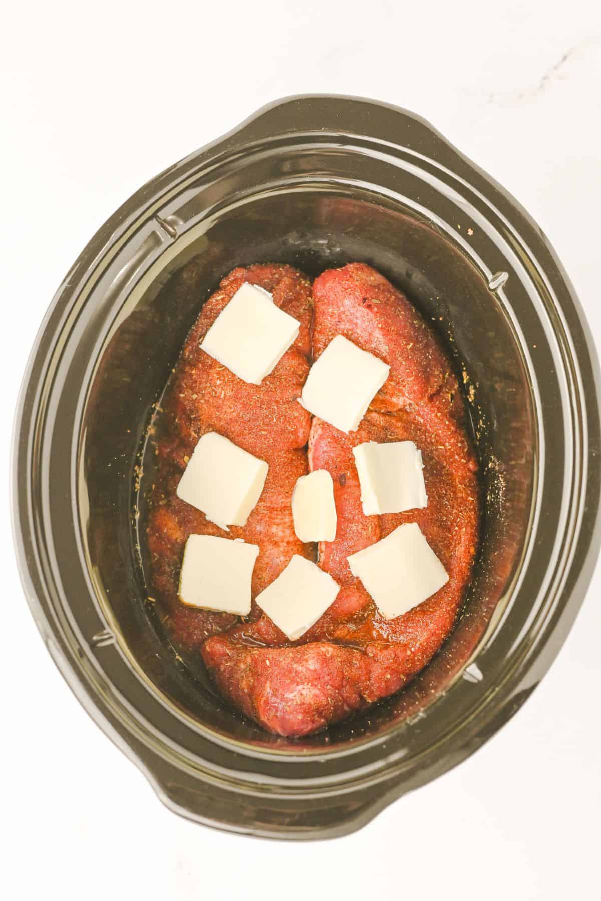 A Crock Pot Pork Tenderloin sits in the slow cooker, featuring two pieces of seasoned meat topped with several small squares of butter, all placed on a white surface.