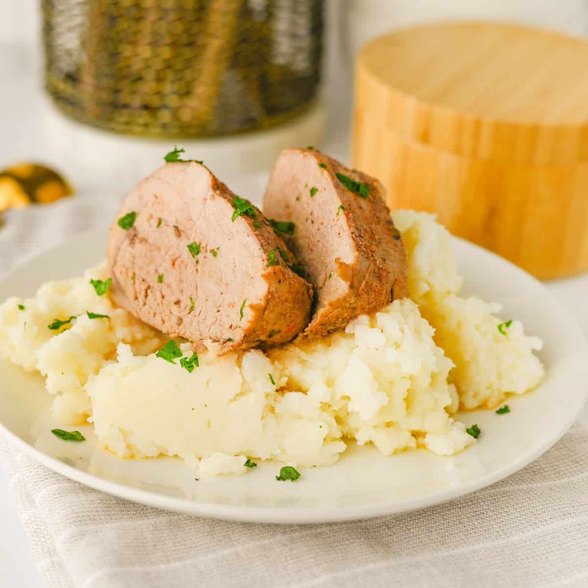 A plate of mashed potatoes topped with two slices of Crock Pot Pork Tenderloin meatloaf, garnished with chopped parsley. The plate sits on a light-colored napkin with a wooden container and a decorative item in the background.