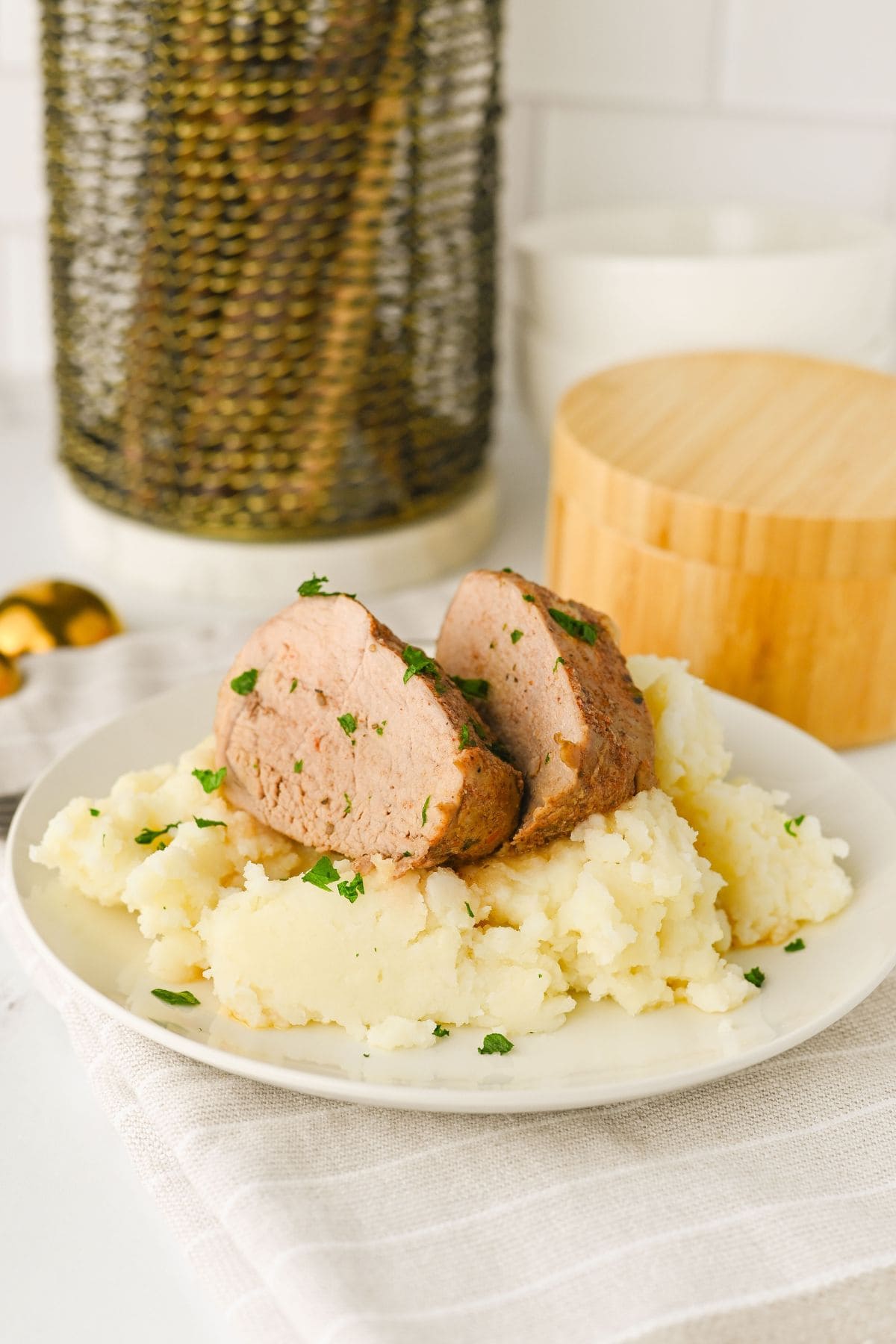 A white plate with mashed potatoes topped with two thick slices of Crock Pot Pork Tenderloin, garnished with chopped herbs. The plate sits on a light-colored cloth with kitchen containers and utensils in the background.
