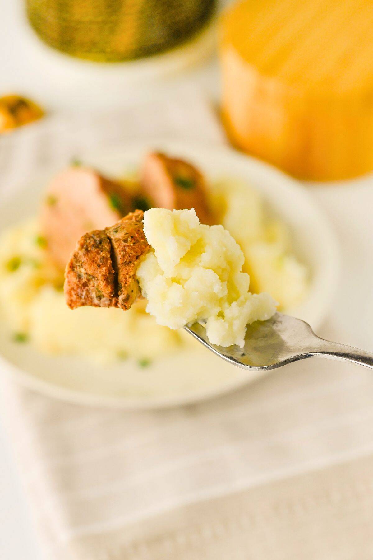 A fork holding a bite of mashed potatoes and Crock Pot Pork Tenderloin, with a plate of more mashed potatoes and tenderloin blurred in the background.