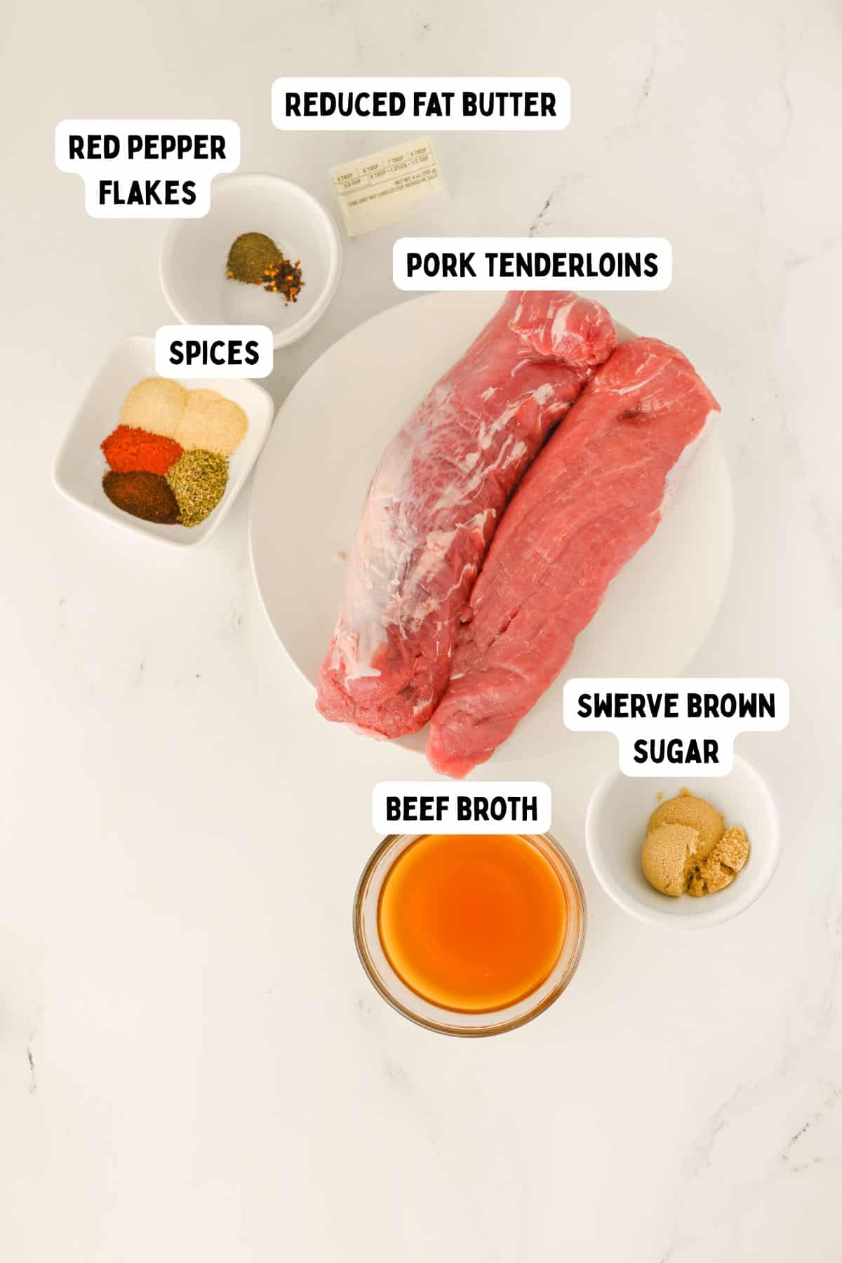 Two raw pork tenderloins on a white plate, surrounded by labeled bowls of spices, red pepper flakes, swerve brown sugar, beef broth, and reduced fat butter—perfect for preparing a flavorful Crock Pot Pork Tenderloin.