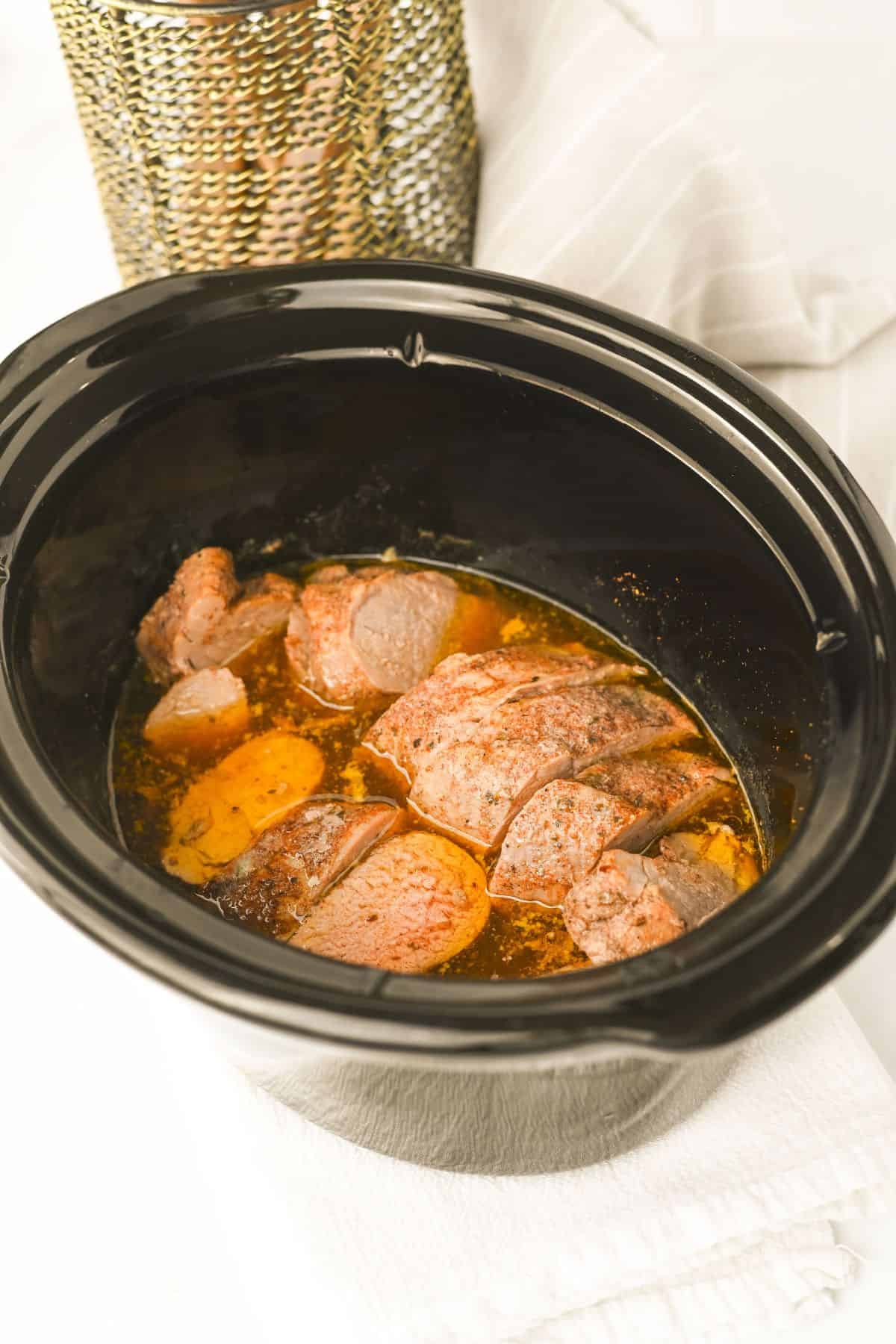 A black slow cooker filled with tender Crock Pot Pork Tenderloin in broth. The cooker rests on a white towel, with a gold textured container and a white cloth in the background.