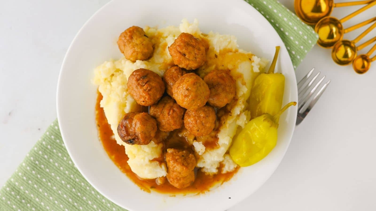 A white plate with mashed potatoes topped with meatballs and brown gravy, served with two yellow peppers. A fork and gold measuring spoons are on a green and white napkin beside the plate.