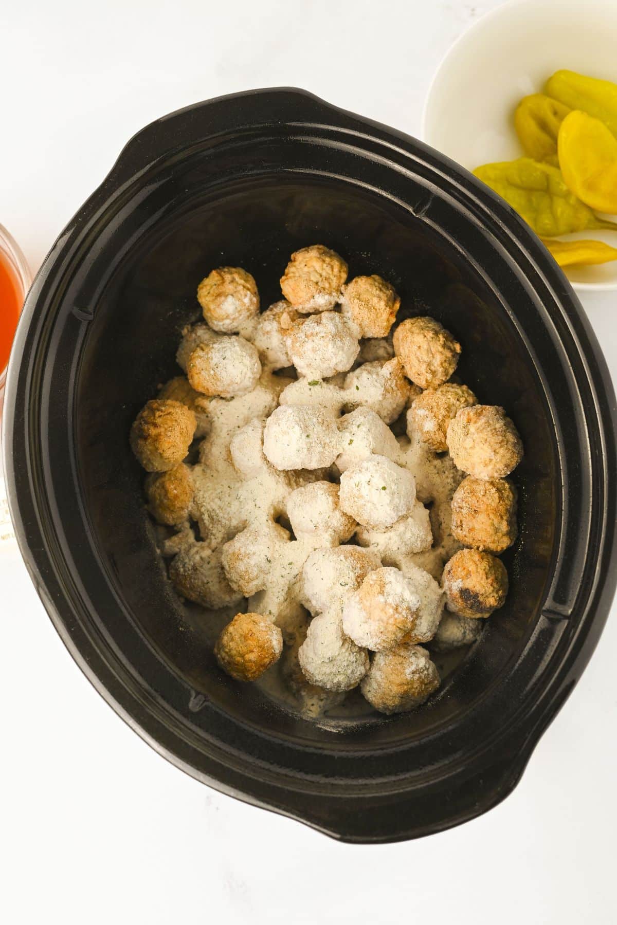 A black slow cooker filled with uncooked meatballs sprinkled with white grated cheese or seasoning. A white bowl with yellow pepper slices is visible to the side on a white surface.