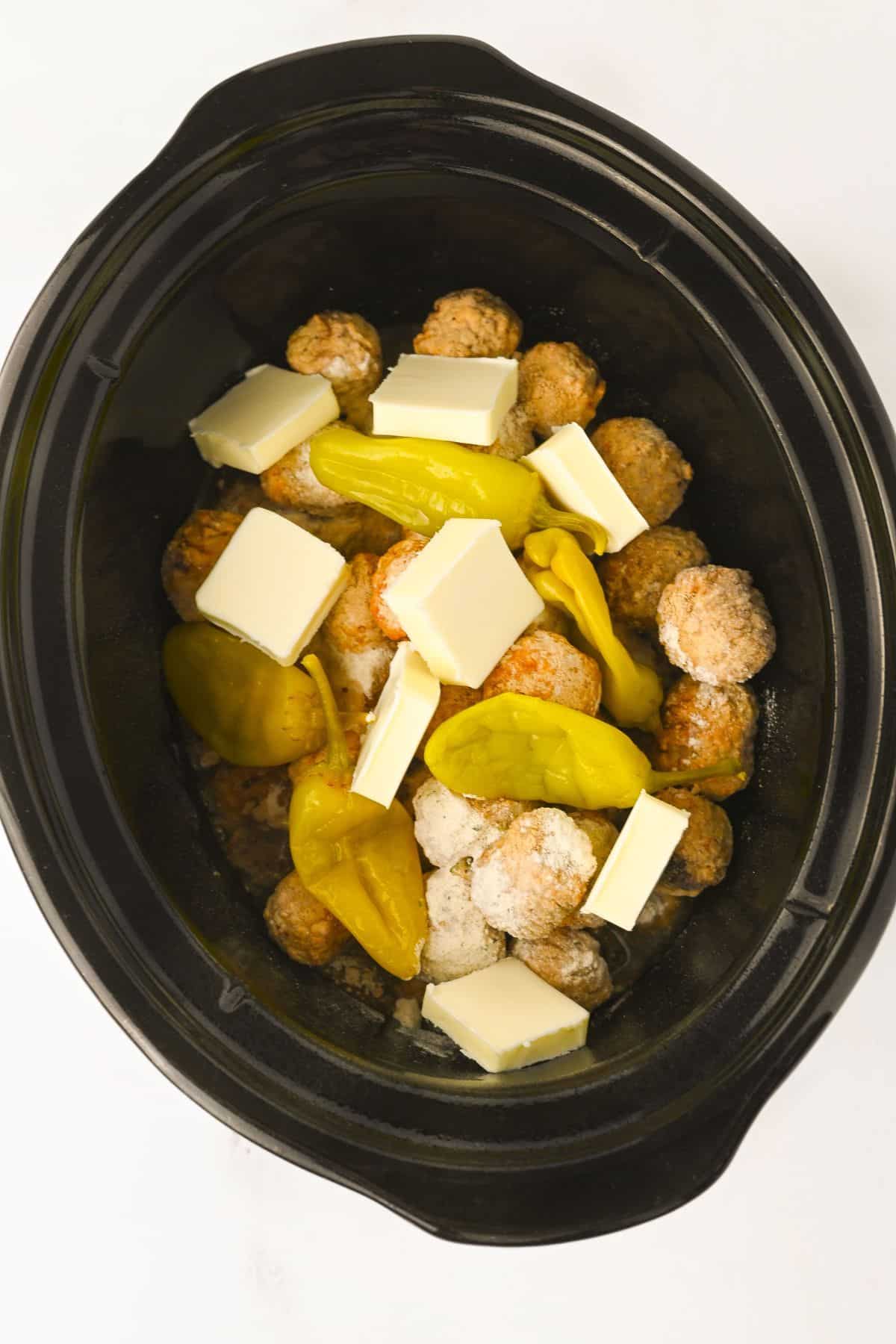 A black slow cooker filled with frozen meatballs, sliced butter, whole yellow pepperoncini peppers, and seasoning on a white surface.