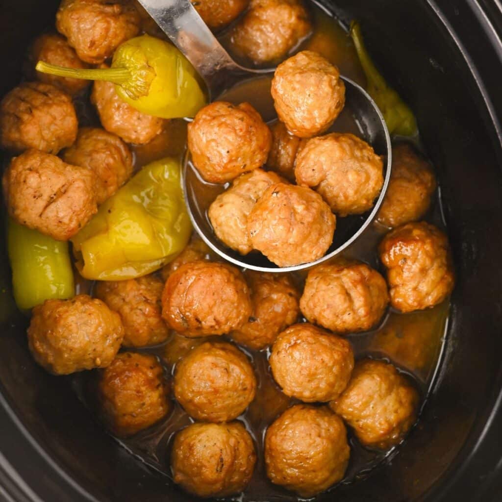 A slow cooker filled with meatballs in sauce, with a metal ladle holding several meatballs above the rest. Whole yellow-green peppers are also visible among the meatballs.