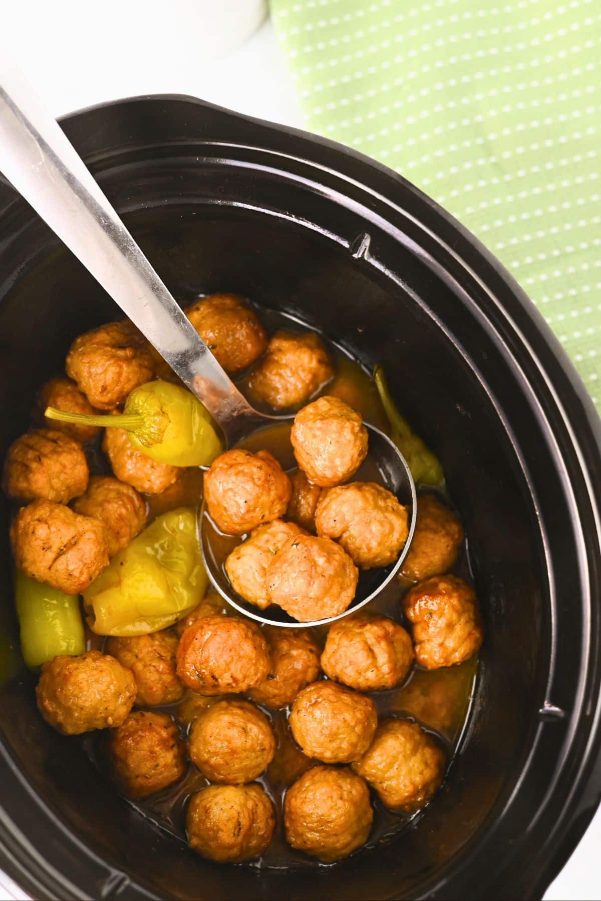 A black slow cooker filled with meatballs, yellow-green peppers, and sauce. A metal ladle is lifting several meatballs from the pot. A green polka dot cloth is partially visible in the background.